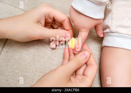 Frau Mutter klebt ein medizinisches Pfandband auf den Kleinkind Baby Finger. Mama s Hand mit einem klebrigen Wundschutzverband und einer Kinderhand. Kind im Alter von einem Ye Stockfoto