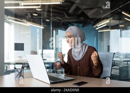 Erfolgreiche Geschäftsfrau Chef feiert Sieg und erfolgreichen Triumph, muslim in Hijab Blick auf Laptop-Bildschirm und hält Hand nach oben Geste des Erfolgs und der Leistung, Frau arbeitet im Inneren Stockfoto