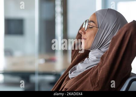 Nahaufnahme verträumte Frau in Hijab und Brille Augen geschlossen mit Händen hinter dem Kopf auf einem Stuhl im Büro sitzend, Business-Frau erfolgreich abgeschlossen ihre Arbeit am Tag ruhen und träumen. Stockfoto