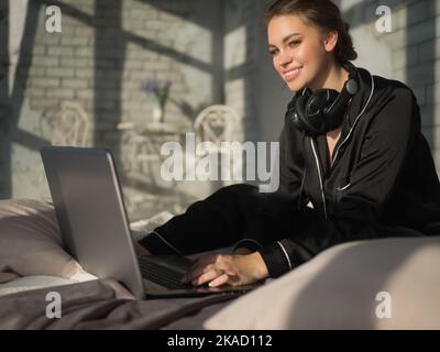 Junge Frau vor ihrem Laptop auf dem Bett zu Hause. Sie schaute auf die Kamera und lächelte. Stockfoto