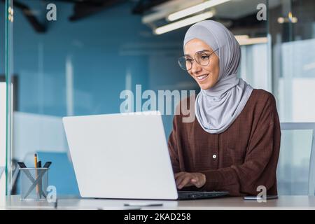 Glückliche und erfolgreiche Geschäftsfrau in Hijab, die mit einem Laptop im modernen Bürogebäude arbeitet, muslimische Frau in einer Brille, die lächelt und glücklich über die Arbeitsleistung ist. Stockfoto