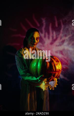 Junge Frau hält Kürbis in den Händen und porträtiert blutrünstigen Zombie mit Horrorwunden im Gesicht und blutigen Kleidern vor dunklem Hintergrund mit Stockfoto