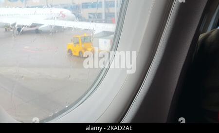 Blick auf den Flughafen durch das Flugzeugfenster, nasses Bullauge, verschwommenes Bild des Flugzeugs, das Konzept des Fliegens bei Regenwetter Stockfoto