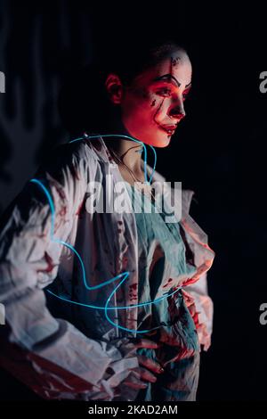 Junge Frau mit leuchtend blauem LED-Band porträtiert blutrünstigen Zombie mit Wunden auf ihrem Gesicht vor dunklem Hintergrund. Gruseliges Bild für Halloween. Stockfoto