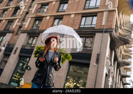 Shooting von unten elegante rothaarige junge Frau trägt Mode Hut mit Smartphone wegschauen stehend mit transparenten Regenschirm von Beautiful Stockfoto