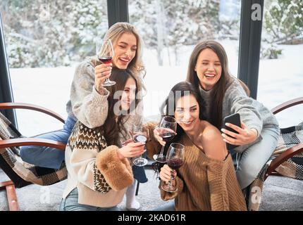 Junge Frauen genießen Winterwochenenden im modernen Scheunenhaus. Vier Mädchen, die Spaß haben, Selfies am Telefon machen und Wein trinken. Stockfoto