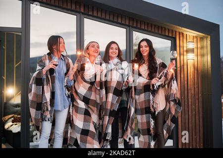 Junge Frauen genießen die Winterwochenenden auf der Terrasse des modernen Barnhouse. Vier Mädchen in Plaids trinken Wein und feiern mit Wunderkerzen am Abend. Stockfoto