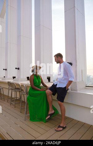 Ein paar Männer und Frauen besuchen ein Dachrestaurant mit Blick auf die Stadt, thailändische Frauen und europäische Männer an einer Dachbar Stockfoto