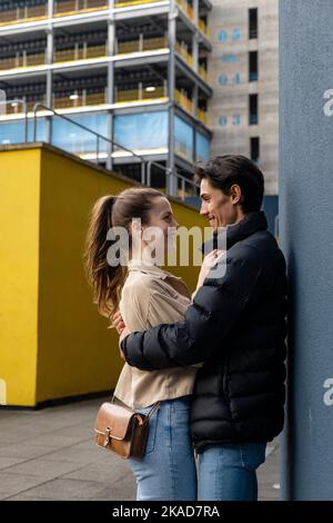 Eine Seitenansicht eines jungen Paares, das von Angesicht zu Angesicht gegen eine Wand steht, sich umarmt und verliebt ist. Stockfoto