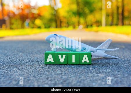 Word AVIA aus Holzblöcken, die vor einem Spielzeugflugzeug im Freien aufgestellt wurden. Günstige Flüge Herbstsaison Konzept. Stockfoto