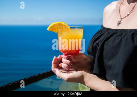 Ein leicht alkoholisches Getränk mit einer Scheibe Orange in den Händen einer Frau vor dem Hintergrund des blauen Meeres. Stockfoto