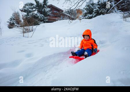 Fröhlicher Junge im orangen Skisport-Outfit geht auf dem Schlitten bergab Stockfoto