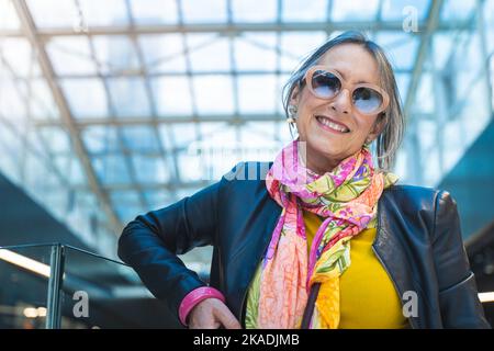Lady mit einem glücklichen Lächeln und Blick auf ein Einkaufszentrum. Ältere Frau mit Sonnenbrillen-Porträt. Stockfoto