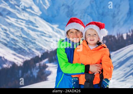 Zwei glückliche Jungen in Weihnachtsmannmützen stehen über Schneebergen Stockfoto