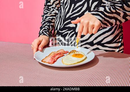 Frau, die Finger in Eigelb steckt. Teller mit englischem Frühstück auf rosafarbener Tischdecke Stockfoto