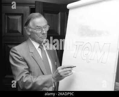 1980s, historisch, in einem Konferenzraum des Hotels, ein geeigneter männlicher Moderator, der neben einem Whiteboard steht und auf dem das Wort „TQM“ steht, steht für „Total Quality Management“, Dies ist ein Managementansatz, der sich auf die Entwicklung langfristiger Geschäftserfolge konzentriert, indem er sich auf den hochwertigen Service jedes einzelnen Mitarbeiters des Unternehmens konzentriert. In dieser Zeit wurden Whiteboards in Präsentationen als visuelle Hilfe verwendet und hatten den Vorteil, dass sie einer Präsentation ein Element der Interaktivität hinzufügen konnten, da Wörter und Kommentare während der Sitzung leicht hinzugefügt oder weggenommen werden konnten. Stockfoto