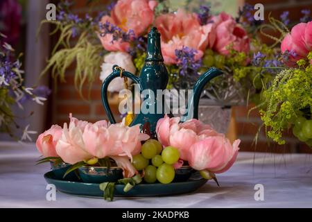 Barocke Blumenarrangements mit pinkfarbenen Pfingstrosen, grünen Trauben und einer orientalischen dunkelgrünen Teekanne auf dem Tisch. Stockfoto