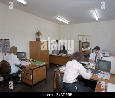 1989, historisches, männliches und weibliches Personal in einem offenen Büro, das an Holztischen sitzt und mit den kleinen Computerterminals der Zeit arbeitet, England, Großbritannien. Stockfoto