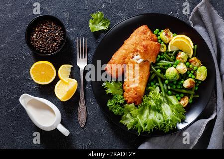 Knuspriger Bierteig Kabeljau Fischfilet mit gebratenem rosenkohl, grünen Bohnen, grünen Erbsen und frischem Salat auf schwarzem Teller auf Betontisch, horizontal Stockfoto