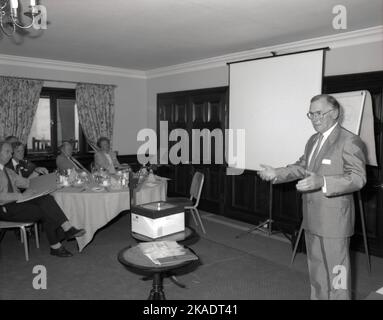 1980s, historisch, in einem Konferenzraum des Hotels, ein geeigneter männlicher Moderator, der neben einem Whiteboard, einer Leinwand und einem Overheadprojektor steht und eine Präsentation vor leitenden Führungskräften des Unternehmens, England, Großbritannien, vorstellt. Der Overheadprojektor oder OHP, wie er genannt wurde, war in dieser Zeit, vor der Einführung des Diaprojektors und der Erstellung von „Audio-visuellen Diashows“, ein Standard-Präsentationstool. Stockfoto