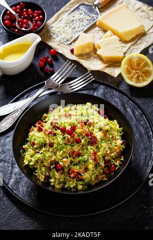 rosenkohl mit Mandeln, knusprig gebratenem Speck und Preiselbeeren in schwarzer Schale auf Betontisch mit Zutaten, vertikale Ansicht von oben Stockfoto