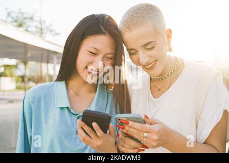 Fröhliche Teenager-Freunde mit dem Handy und lachten in der modernen Stadtstraße. Junge multiethnische Frauen. Stockfoto