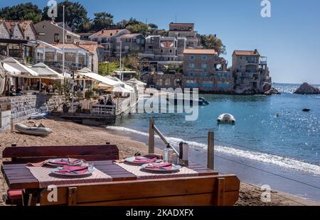 Budva, Montenegro - 28. April 2022: Malerische kleine Bucht am Przno Plaza. Sandstrand, mittelalterliche Stadtarchitektur. Restauranttisch aus Holz im Vordergrund. Stockfoto