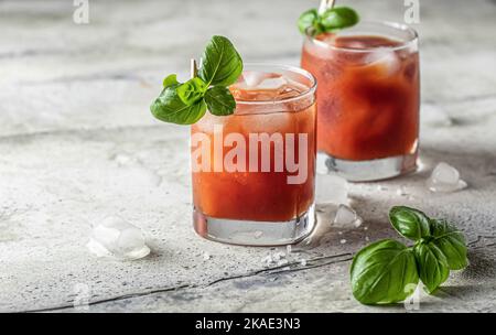 Tomatensaft oder Bloody Mary Eiscocktail mit Bazilblättern in zwei Gläsern für Getränkekarte auf grauem texturierten Hintergrund mit Textraum Stockfoto