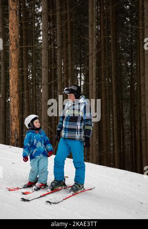 Der kleine Junge lernt mit seinem Vater Ski zu fahren oder steht auf einem schneebedeckten Berghang vor dem Hintergrund von Tannenbäumen. Ein interessanter Aktivist Stockfoto