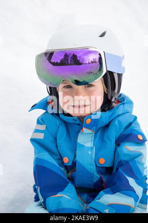 Portrait Kind Junge in einem Helm, Skimaske und warme Jacke sitzt auf dem Schnee. Winterspaß für Erwachsene und Kinder. Der kleine Junge lernt im Winter Skifahren Stockfoto