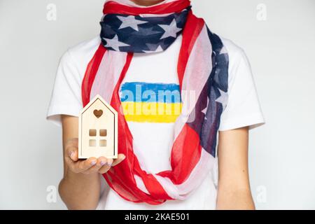 Ein Mädchen mit einer amerikanischen Flagge um den Hals mit einer ukrainischen Flagge auf ihrem T-Shirt hält ein kleines Holzhaus in der Hand auf weißem Hintergrund, Leben Stockfoto