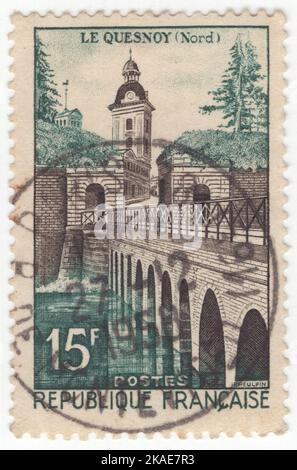 FRANKREICH - 1957. Juli 19: Eine dunkelblau-grüne und sepiafarbene Briefmarke mit 15 Franken, die Le Quesnoy, Gemeinde und kleine Stadt im Osten des Nordfrankreichs darstellt. Es war Teil der historischen Provinz des französischen Hennegaus. Vor Ende des Jahres 1940s hatte das Unternehmen eine wichtige Branche im Schuhmacherbereich, gefolgt von einer Chemiefabrik und einer Molkerei, die dem Wochenmarkt, dem Tourismus und dem Pendeln zu anderen Orten wie Valenciennes und lokalen Geschäften Platz eingab Stockfoto