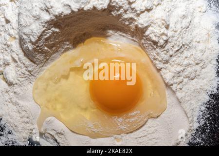 HühnerEigelb auf Mehl in Nahaufnahme. Zerbrochenes Ei auf Mehl, bedeutet für die Herstellung von Brot. Der Prozess der Herstellung von Teig für Kekse, Pasta, Brot, Kuchen. Hausmannskost. Stockfoto