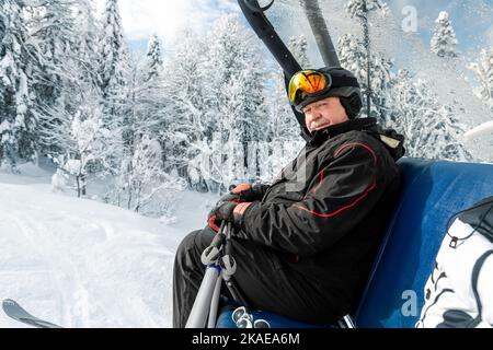 Aktiver älterer erfahrener älterer älterer Mensch in Skihelm, Schutzbrille und schwarzem Anzug sitzen auf dem Skilift Berggipfel ruhen Genießen Sie kalten Extremsport Stockfoto