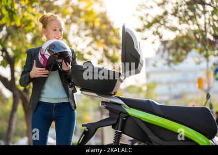 Eine rothaarige Frau, die ihren Helm im Kofferraum ihres geparkten Motorrads hält Stockfoto