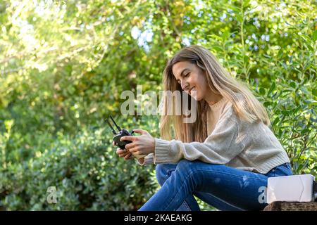 Low-Angle-Ansicht einer jungen blonden Frau, die lächelt und glücklich ist, während sie mit einer Fernbedienung in den Händen eine Drohne fliegt Stockfoto