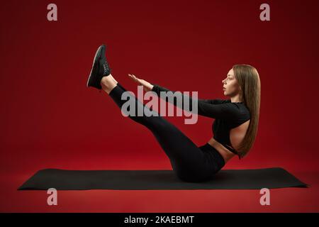 Seitenansicht einer weiblichen Trainerin in einer schwarzen Suite, die mit einem Übungsboot posiert, um die Beine hochhalten zu können. Brunette Fitness-Trainer zeigt, wie Affektivität Aktivität für beste Sportform. Stockfoto