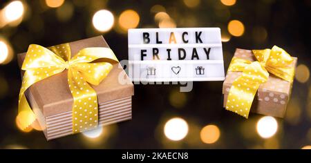 Kreatives Werbebanner mit Geschenkboxen mit gelben Polka-Dot-Bändern und Schleifen sowie weißem Leuchtkasten mit Black Friday-Text auf Schwarz mit goldenem Bokeh Stockfoto