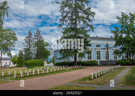 Finnland, Hamina - 18. Juli 2022: Kriegsfriedhof des Zweiten Weltkriegs mit Reihen von weißen Kreuzen und roten Blumen auf Rasen neben der blauen St. Johns Kirche, Johanneksen k Stockfoto