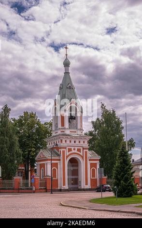 Finnland, Hamina - 18. Juli 2022: Rot-weißer Glockenturm der Peter-und-Paul-Kirche und Eingang unter einer schweren grauen Wolkenlandschaft mit grünem Laub. Autos in s Stockfoto