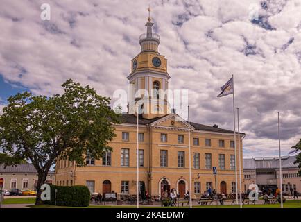 Finnland, Hamina - 18. Juli 2022: Stadt oder Rathaus aus gelbem Stein, Haminan raatihuone, mit Uhrenturm unter der grauen Wolkenlandschaft. Grüner Rasen und Peop Stockfoto