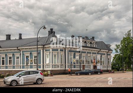 Finnland, Hamina - 18. Juli 2022: Historisches blau-weißes Geschäftsgebäude gegenüber dem Rathaus unter grauer Wolkenlandschaft mit Autos und Menschen davor. Cultu Stockfoto