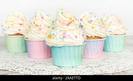 Cupcakes aus Vanille mit Buttercreme, garniert mit essbaren goldenen Streuseln aus nächster Nähe. Muffins in bunten Pappbechern stehen auf einer weißen Spitzenserviette auf einer fe Stockfoto
