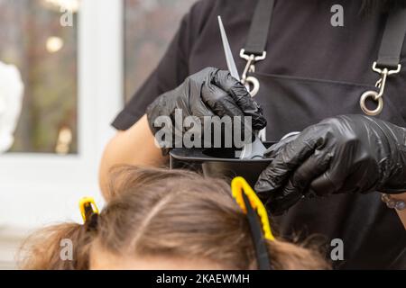 Mädchen in schwarzen Gummischutzhandschuhen, die eine Bürste in den Händen halten und Haarfärbemittel aus der Nähe rühren. Colorist Haarpflege im Schönheitssalon Haarmaske. Stockfoto