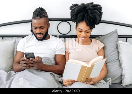 Glückliche Ehegatten, afroamerikanischer Mann und Frau, sitzen im Bett im Schlafzimmer, der Mann benutzt ein Smartphone, liest die Nachrichten, stöbert im Internet, die Frau liest ein Interessantes Buch, bevor sie zu Bett geht Stockfoto