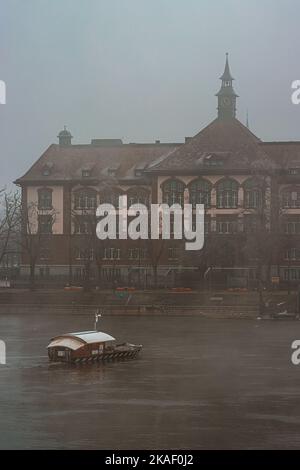 Eine vertikale Aufnahme eines Hausbootes im Rhein mit einem Küstengebäude am nebligen Morgen, Basel, Schweiz Stockfoto