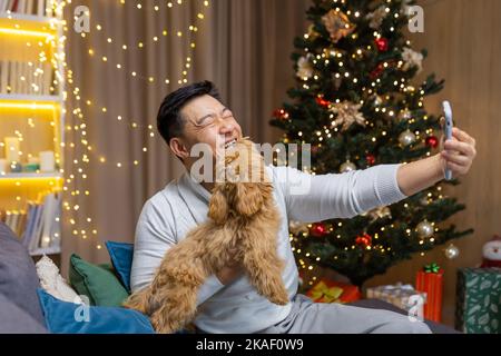 Junger glücklicher, gutaussehender asiatischer Mann hält Telefon und nimmt Selfie mit seinem Hund. Am Weihnachtsbaum zu Hause sitzen. Stockfoto