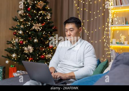 Ein junger asiatischer Geschäftsmann arbeitet im Urlaub an einem Laptop. Er sitzt ernst zu Hause auf dem Sofa in der Nähe des Weihnachtsbaums. Stockfoto