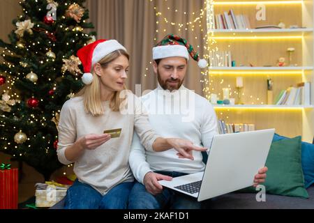 Festliche Online-Einkäufe für die ganze Familie. Ein junges Paar, ein Mann und eine Frau, sitzen zu Hause auf dem Sofa in der Nähe des Weihnachtsbaums in roten Hüten, halten einen Laptop und eine Kreditkarte. Stockfoto