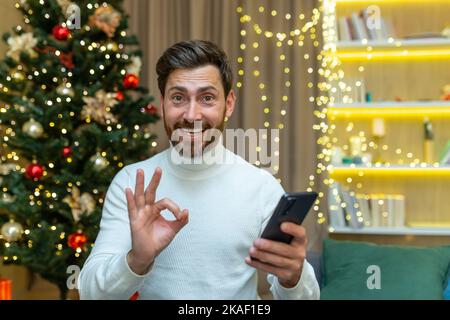 Festliches Online-Shopping. Ein junger Mann hält ein Telefon in den Händen, kauft online ein, kauft Geschenke zu Weihnachtsrabatten, zum Verkauf. Er blickt auf die Kamera, lächelt, zeigt mit den Fingern. Stockfoto